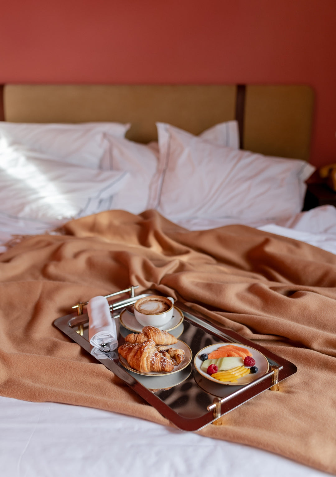 TBCo Camel Lambswool Blanket draped on white bedding with breakfast tray featuring croissant, coffee and fresh fruit